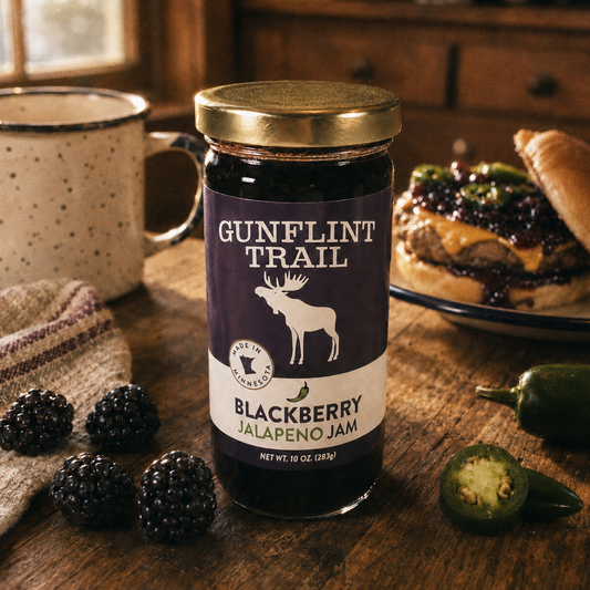 A jar of Gunflint Trail Blackberry Jalapeño Jam sits on a rustic wooden table, surrounded by fresh blackberries, a sliced jalapeño, a sandwich with jam, and a speckled mug in a warm kitchen setting.