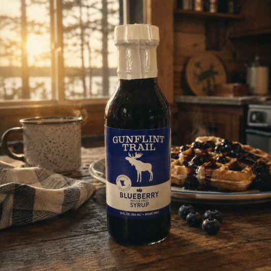 A bottle of Gunflint Trail Blueberry Syrup stands on a rustic wooden table in a warm kitchen setting, with a plate of blueberry waffles, blueberries, and a mug in the background.