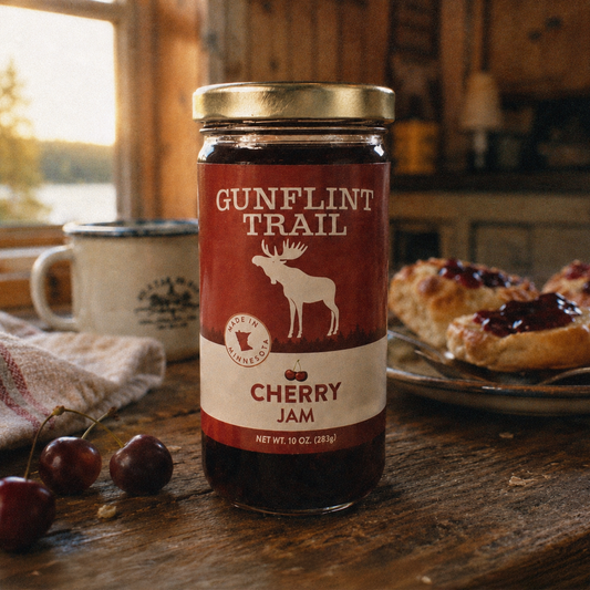 A jar of Gunflint Trail Cherry Jam stands on a rustic wooden table in a cozy cabin setting, with fresh cherries, a mug, and a plate of berry-topped pastries nearby.