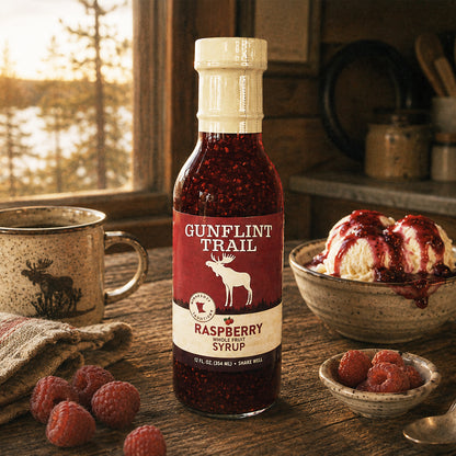 A bottle of Gunflint Trail Raspberry Syrup stands on a rustic wooden table in a cozy kitchen setting, surrounded by fresh raspberries, a mug, and a bowl of ice cream with syrup drizzle. The label clearly shows the brand name 'Gunflint Trail'.