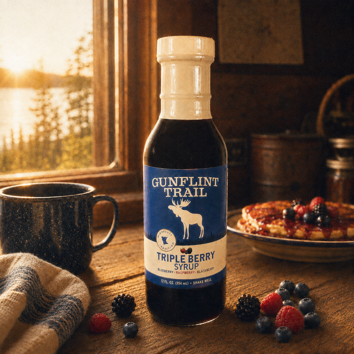 A bottle of Gunflint Trail Triple Berry Syrup stands on a rustic wooden table in a cozy kitchen setting. The bottle has a blue label with a white bear logo and is surrounded by berries, a dark mug, and pancakes with fruit on a plate in the background, with warm sunlight streaming through a window.